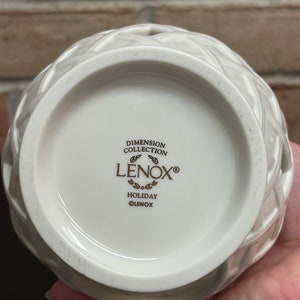 May include: A white ceramic Lenox Holiday Dimension Collection vase with a woven design. The bottom of the vase features the Lenox logo and the words "Dimension Collection" and "Holiday".