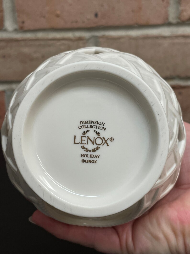 May include: A white ceramic Lenox Holiday Dimension Collection vase with a woven design. The bottom of the vase features the Lenox logo and the words "Dimension Collection" and "Holiday".