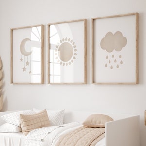 May include: Three framed prints with minimalist illustrations of a crescent moon with stars, a sun, and a cloud with raindrops. The prints are hung on a white wall above a white bed with beige bedding.