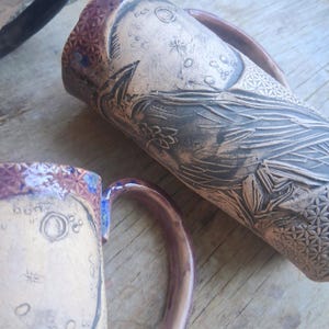May include: Two handmade ceramic mugs. One mug features a detailed raven design, celestial elements, and floral patterns. The other mug has a similar design with a purple rim and handle. Both mugs are in earthy tones.