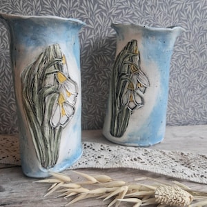 May include: Two handmade ceramic vases with a light blue and white glazed finish. Each vase features a raised, sculpted relief of white snowdrop flowers with green stems and yellow centres. The vessels have an irregular, wavy rim and a smooth texture.