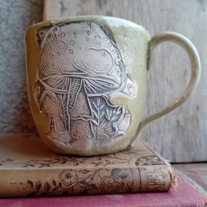 May include: A handcrafted ceramic mug with a pale green glaze, featuring an etched mushroom design. The mushroom is detailed with a contrasting colour, showing the cap, stem, and surrounding plants. The mug has a curved handle and rests on a vintage book.