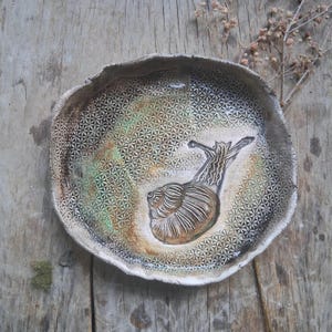 May include: Handmade ceramic plate with a textured surface and a raised snail design. The plate features a geometric pattern and earth-tone colours, including brown, green, and beige. The plate is set on a wooden surface.