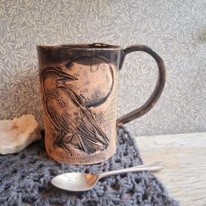 May include: A handcrafted ceramic mug with a raven and moon design. The mug is brown with a dark handle and rim. The raven is carved into the side of the mug, with a moon in the background. A silver spoon rests on a gray crocheted coaster.