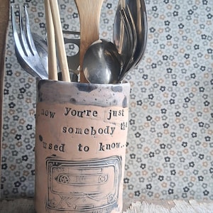 May include: A ceramic utensil holder with the text "Now You're just somebody that used to know..." and a cassette tape design. The holder contains wooden spoons, chopsticks, and metal forks and spoons. The holder is beige with a speckled rim.