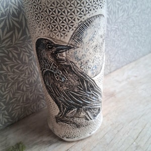 May include: A cream ceramic vase featuring a detailed raven illustration. The raven is black and grey, set against a moon design. The vase has a geometric pattern and is approximately 20 cm tall.