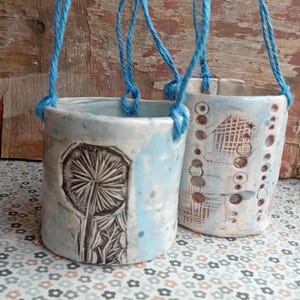 May include: Two handmade ceramic hanging planters. The planters are cylindrical with a light blue and white glaze. One features a black etched dandelion design, the other has a pattern of circles and lines. They hang from blue twine.