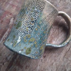 May include: A handcrafted ceramic mug with a distinctive design. The mug has a textured pattern of small, repeating floral shapes. The colour transitions from a light beige at the top to a teal blue at the bottom, with green and yellow accents.