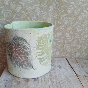 May include: A handmade ceramic cylindrical vase with a light green interior and exterior. The exterior features leaf designs in shades of green and pink. The vase is speckled with black dots and sits on a wooden surface.