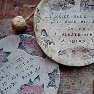 May include: Two handcrafted ceramic plates with floral and mushroom patterns. One plate has the inscription "YOU ARE THE ONLY THING IN ANY ROOM YOU'RE EVER IN." The other plate reads "I NEVER MAKE THE SAME MISTAKE TWICE I ALWAYS AIM A THIRD TI."