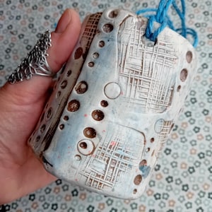 May include: A handmade ceramic pendant with a textured surface, in white, light blue, and brown. It features circular and linear patterns, with a blue cord for hanging. The pendant is held in a hand. The pendant is approximately 7 cm in length.