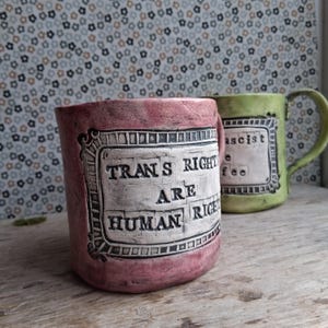 May include: Two ceramic mugs with a distressed finish. One mug is pink and says "Trans Rights Are Human Rights" in a white banner. The other mug is green and says "Fascist" in a white banner.