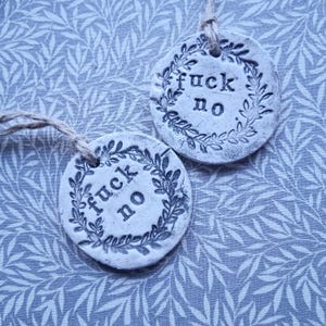 May include: Two round, off-white ceramic ornaments with the words "fuck no" in a dark gray font. Each ornament is encircled by a wreath of leaves and has a twine loop for hanging. The background is a light blue fabric with a leaf pattern.