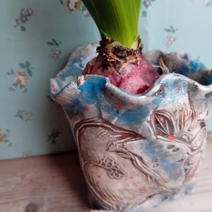 May include: A handmade ceramic plant pot with a blue and white design, featuring a bird motif. The pot holds a bulb with green leaves sprouting. The pot has a wavy rim and is decorated with red speckles.