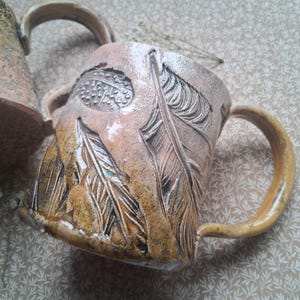 May include: Handmade ceramic mug with a textured surface and feather design. The mug features a tan and brown colour palette with a curved handle. The feathers are carved into the surface, adding a unique, artistic touch. The mug is ideal for coffee or tea.