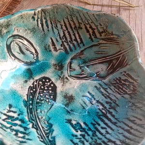 May include: A handcrafted ceramic bowl with a unique, textured surface. The bowl features a turquoise and black colour scheme with abstract feather and leaf designs. The rim has an irregular, wavy shape. The bowl is ideal for decorative purposes.