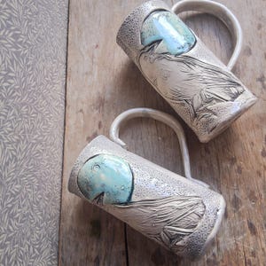 May include: Two ceramic mugs with a light gray base and a detailed bird design. Each mug features a turquoise blue accent and a curved handle. The mugs are placed on a wooden surface.