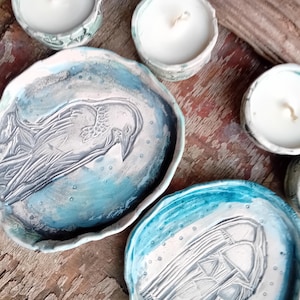 May include: Handmade ceramic raven-themed trays and tealight candle holders. The trays are painted in shades of blue and white, with a raised raven design. The tealight holders are small, round, and also painted in blue and white. The items are arranged on a wooden surface.