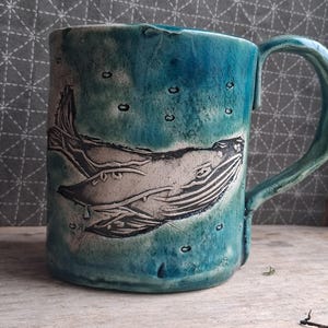 May include: A handmade ceramic mug with a whale design. The mug has a turquoise glaze with a whale illustration in white and black. The mug has a handle and is suitable for hot or cold drinks.