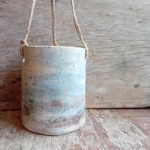 May include: A handmade ceramic hanging planter, cylindrical in shape, with a blue, white, and brown speckled design. It is suspended by three natural fibre ropes. The planter is ideal for small plants. The rustic design adds a touch of nature to any space.