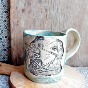 May include: A ceramic mug with a blue and green glaze. The mug features a black and white illustration of a twenty-sided die.
