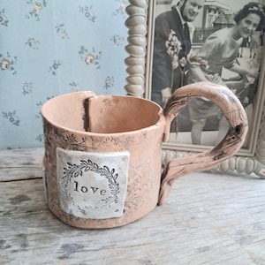 May include: A light peach-colored ceramic mug with a textured surface and a curved, branch-like handle. A square white plaque is attached to the front, featuring a stamped laurel wreath design surrounding the lowercase word "love" in black ink.