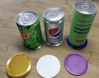 May include: Three aluminum beverage cans with different labels and colors, sitting on colorful round coasters. The cans are on a wooden surface. The labels read "Diet Pepsi", "Mountain Dew", and "Trader Joe's Sparkling Watermelon Juice".