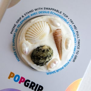 May include: A PopGrip phone grip and stand with a swappable top. The round design features seashells, pearls, and a small rock set in a white base. The packaging includes the text "Phone Grip & Stand" and "Try Me! Pull Twice to Grip."