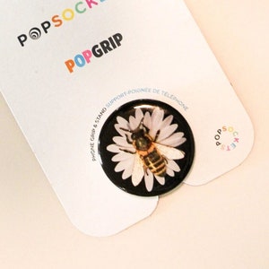 Puede incluir: Una abeja blanca y amarilla sobre una margarita blanca sobre un fondo negro. La imagen está en una tarjeta blanca con el texto "PopSockets PopGrip" en letras de colores.