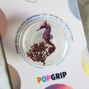 May include: A purple seahorse with a brown seaweed design inside a clear plastic pop grip. The pop grip is attached to a white background with the text "POPGRIP" in pink, blue, and black.