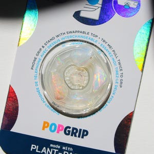 Könnte beinhalten: Ein klarer, schillernder PopGrip-Telefonhalter mit austauschbarem Aufsatz, hergestellt aus pflanzlichen Materialien. Die Verpackung enthält Anweisungen auf Englisch und Französisch. Der Griff ist rund mit einem klaren, strukturierten Zentrum und holografischen Akzenten.