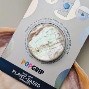 May include: A circular phone grip and stand with an iridescent abalone shell finish, mounted on a grey retail card. The packaging features the text POPSOCKETS, POPGRIP, and MADE WITH PLANT-BASED MATERIALS, along with diagrams showing usage.