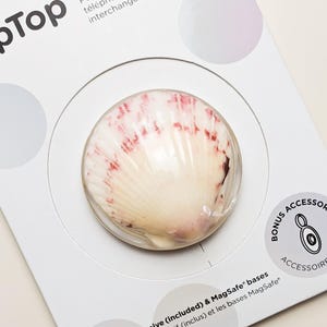 Può includere: Un supporto e impugnatura circolare per telefono con un design a conchiglia realistico nei toni del bianco e del rosa rossastro. Il prodotto è montato su una confezione bianca con il testo Phone grip & stand with swappable top + adhesive base.