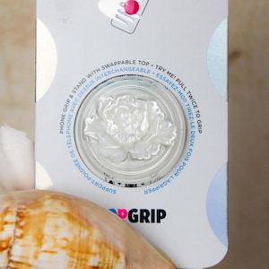 May include: White phone grip with a pearl-like flower design. The grip is round and has a raised edge. The text "GRIP" is printed in black on the bottom of the grip. The text "PHONE GRIP & STAND WITH SWAPPABLE TOP TRY ME! PULL TWICE TO GRIP" is printed on the white background.