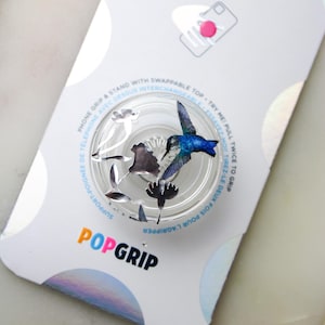 May include: A clear phone grip with a hummingbird and floral design. The grip is expandable and has a swappable top. The packaging says "PopSockets PopGrip".