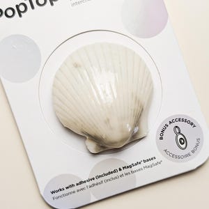 Può includere: Un supporto e impugnatura per telefono a forma di conchiglia bianca e beige su una scheda di vendita bianca. La confezione riporta il testo PopSockets e PopTop, indicando che si tratta di un supporto con parte superiore intercambiabile.