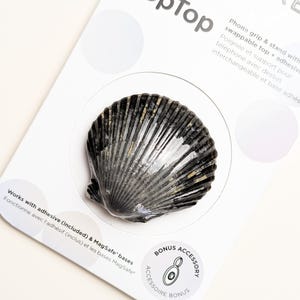 Può includere: Un supporto e impugnatura per telefono a forma di conchiglia nera e grigia su una confezione bianca. La confezione riporta il testo PopSocket e PopTop, informazioni sulla compatibilità con adesivo e MagSafe e un'icona di accessorio bonus.