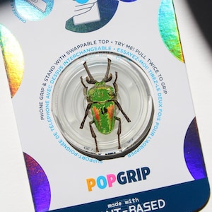 May include: A circular phone grip and stand featuring a green and red beetle design on a clear base. The packaging is white with holographic accents and includes the text POPGRIP, made with PLANT-BASED materials, and instructions to pull twice to grip.
