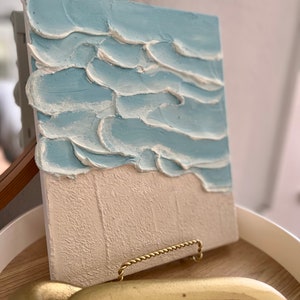 Op de afbeelding: Getextureerd canvas kunstwerk dat een strandsc&egrave;ne uitbeeldt. Het bovenste gedeelte toont lichtblauwe golven met witte accenten, terwijl de onderste helft een beige zandstrand laat zien. Het kunstwerk wordt gepresenteerd op een houten dienblad met een gouden handvat.