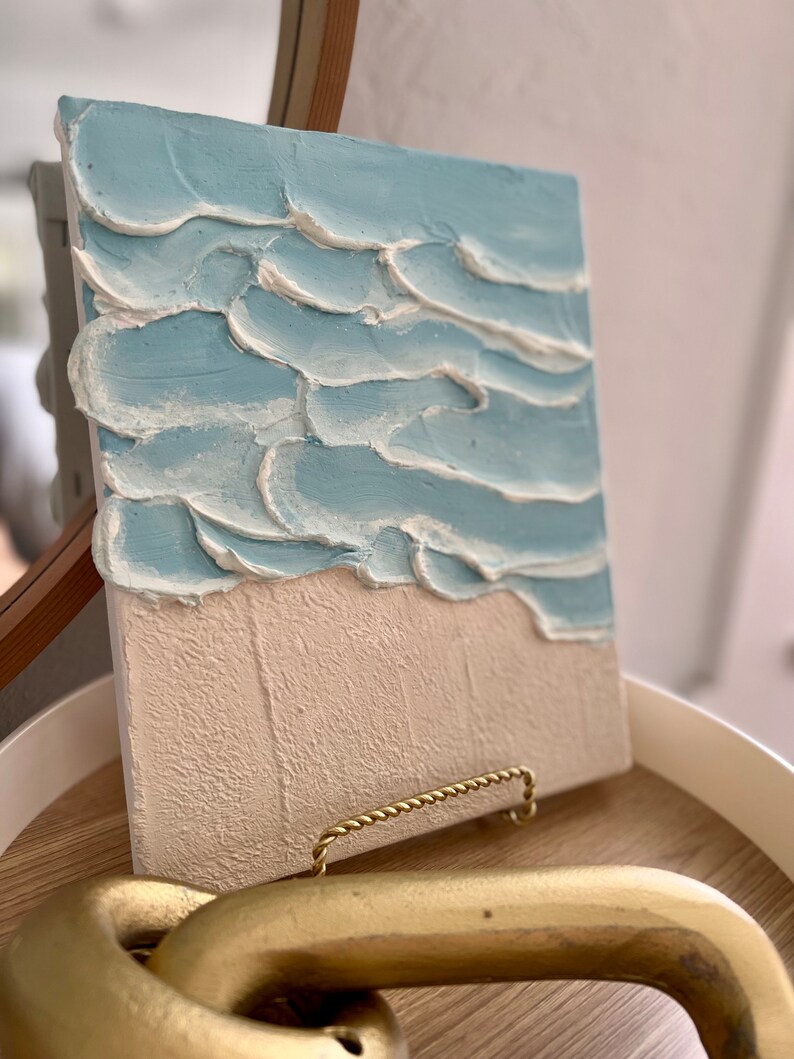 Op de afbeelding: Getextureerd canvas kunstwerk dat een strandsc&egrave;ne uitbeeldt. Het bovenste gedeelte toont lichtblauwe golven met witte accenten, terwijl de onderste helft een beige zandstrand laat zien. Het kunstwerk wordt gepresenteerd op een houten dienblad met een gouden handvat.