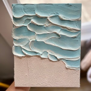 Op de afbeelding: Getextureerd canvas kunstwerk dat een strandsc&egrave;ne uitbeeldt. Het kunstwerk toont een lichtblauwe oceaan met witte accenten en een getextureerd, gebroken wit zandgebied. De verf is aangebracht in dikke, gelaagde streken, waardoor een 3D-effect ontstaat. Het canvas is rechthoekig.