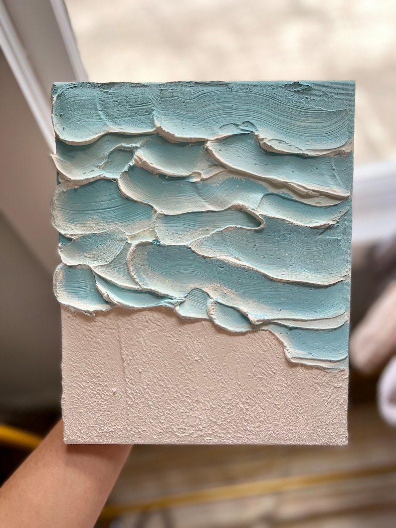 Op de afbeelding: Getextureerd canvas kunstwerk dat een strandsc&egrave;ne uitbeeldt. Het kunstwerk toont een lichtblauwe oceaan met witte accenten en een getextureerd, gebroken wit zandgebied. De verf is aangebracht in dikke, gelaagde streken, waardoor een 3D-effect ontstaat. Het canvas is rechthoekig.