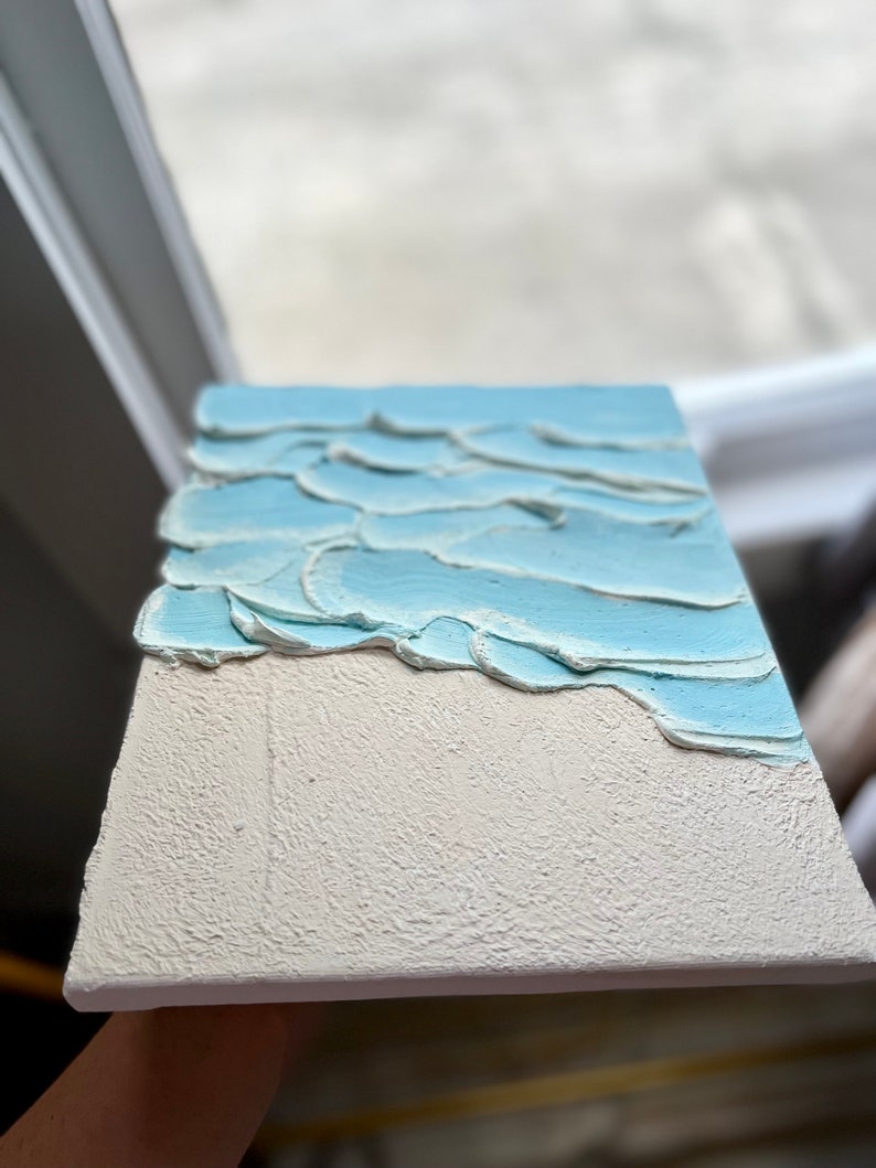 Op de afbeelding: Getextureerde canvas kunst die een strandsc&egrave;ne uitbeeldt. Het onderste gedeelte is getextureerd wit, wat zand voorstelt. Het bovenste gedeelte heeft lichtblauwe, getextureerde golven. Het kunstwerk is rechthoekig en lijkt handgemaakt.