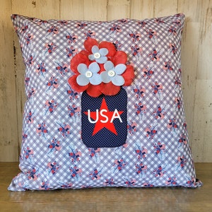 May include: A decorative pillow with a gingham pattern in shades of blue and white. The pillow features a red, white, and blue applique of a mason jar with flowers and the word "USA" on the front.