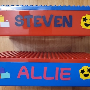 May include: Two red and blue plastic storage boxes with Lego-style lids. The top box has the name "STEVEN" and a yellow smiley face with stars for eyes. The bottom box has the name "ALLIE" and a yellow smiley face with stars for eyes.