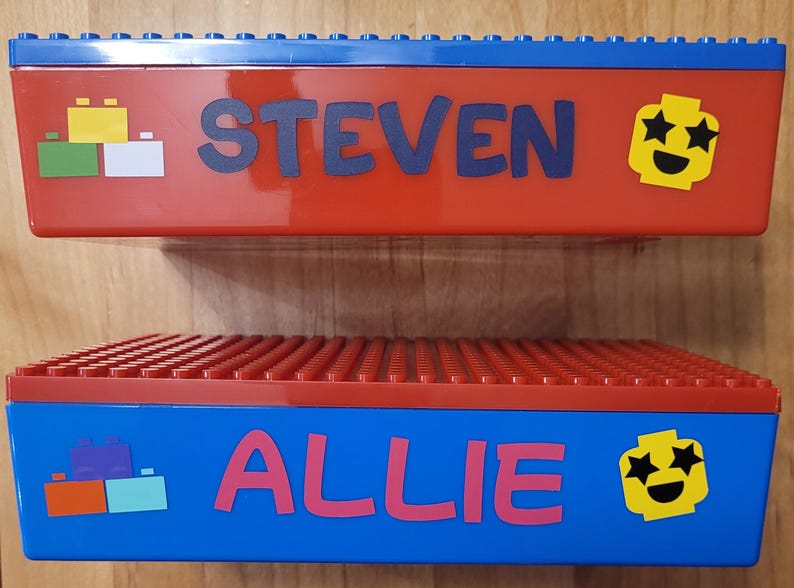 May include: Two red and blue plastic storage boxes with Lego-style lids. The top box has the name "STEVEN" and a yellow smiley face with stars for eyes. The bottom box has the name "ALLIE" and a yellow smiley face with stars for eyes.