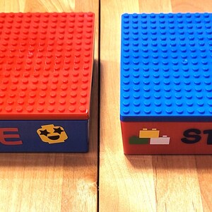 May include: Two Lego storage boxes, one red and one blue, with personalized names "Allie" and "Steven" on the front. Each box has a smiley face sticker and a small Lego brick design.