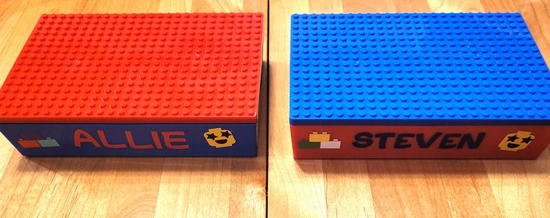 May include: Two Lego storage boxes, one red and one blue, with personalized names "Allie" and "Steven" on the front. Each box has a smiley face sticker and a small Lego brick design.