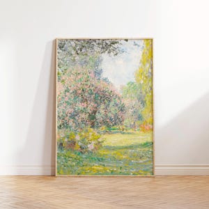Op de afbeelding: Een ingelijst landschapsschilderij met een lichte houten lijst. Het kunstwerk toont een tuinscène met roze bloeiende bomen, groen gebladerte en een grasveld. Het schilderij heeft een zachte, impressionistische stijl met focus op licht en kleur.