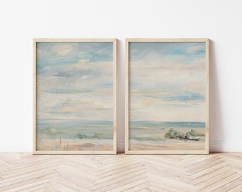 May include: Two framed landscape paintings. Each features a serene beach scene with a light blue sky, soft clouds, and a sandy shore. The artwork is in a muted color palette, with beige wooden frames.